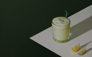green frothy drink in a glass sitting on a white table surface next to a slice of lemon and a slice of lime